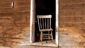 Old, chipped white wooden chair on a weathered wooden porch inside an open doorway of a rustic shed.