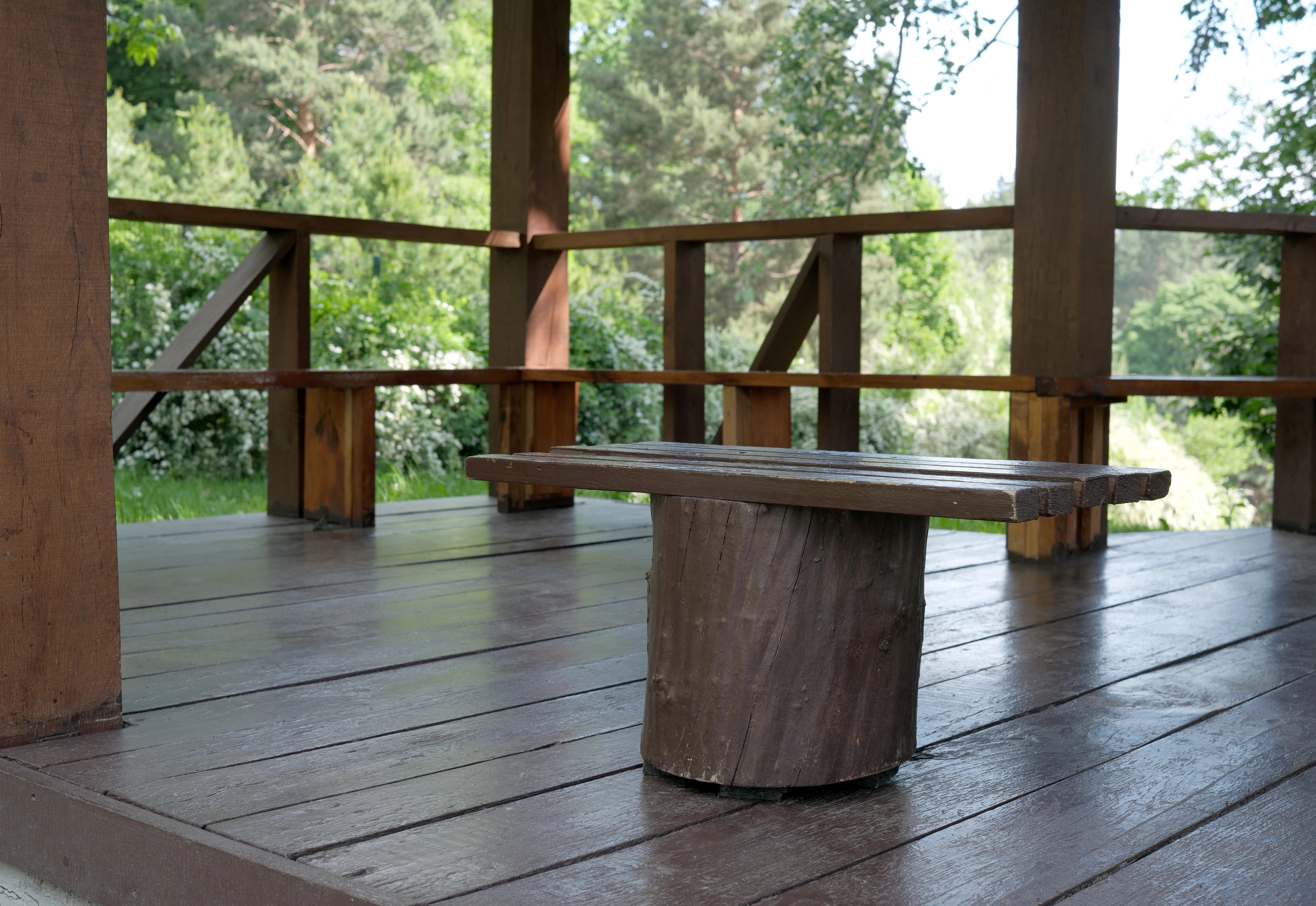 Wooden gazebo/deck with thick posts and railing, a wooden bench in the foreground, and lush green trees in the background.